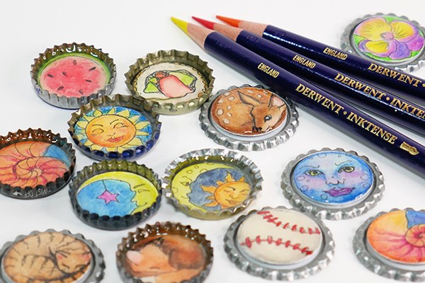 Bottle caps with intricate paintings on them laying next to Derwent Inktense pencils